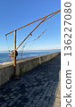A weathered crane structure stands next to a seaside promenade, casting shadows under a clear blue sky.  136227080