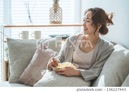 Smiling Asian woman sitting on the sofa in the living room and eating popcorn Smiling Asian woman sitting on the sofa in the living room and eating popcorn 136227443