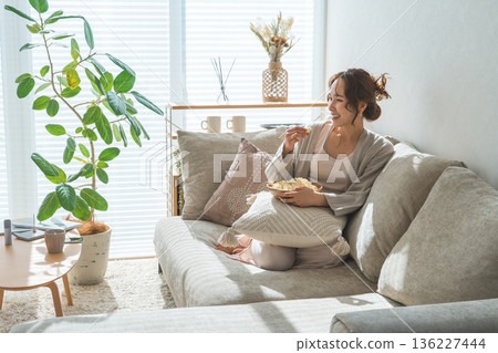 一位微笑的亞洲女性坐在客廳的沙發上吃爆米花。 一位微笑的亞洲女性坐在客廳的沙發上吃爆米花。 136227444