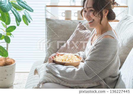 一位微笑的亞洲女性坐在客廳的沙發上吃爆米花。 一位微笑的亞洲女性坐在客廳的沙發上吃爆米花。 136227445