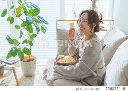 一位微笑的亞洲女性坐在客廳的沙發上吃爆米花。 一位微笑的亞洲女性坐在客廳的沙發上吃爆米花。 136227446