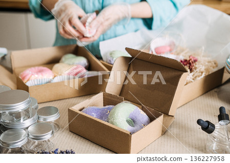 Person places colorful handmade soap products into cardboard boxes with paper filler and clear jars. Handmade bath bombs, personalized self-care gifts, bath salt mixology, aesthetic gift packaging. Person places colorful handmade soap products into cardboard boxes with paper filler and clear jars. Handmade bath bombs, personalized self-care gifts, bath salt mixology, aesthetic gift packaging. 136227958