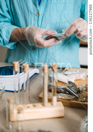 Hands in gloves pour dark minerals from small packet into glass test tube on digital scale. Waterless concentrate, anhydrous DIY, preservative-free bath, solid mineral skincare. Hands in gloves pour dark minerals from small packet into glass test tube on digital scale. Waterless concentrate, anhydrous DIY, preservative-free bath, solid mineral skincare. 136227966