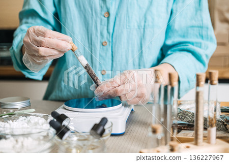Gloved hands hold glass test tube filled with salt and botanical layers near digital scale. Anhydrous skincare, solid bath salts, waterless concentrate, botanical layering. Gloved hands hold glass test tube filled with salt and botanical layers near digital scale. Anhydrous skincare, solid bath salts, waterless concentrate, botanical layering. 136227967