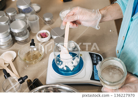 Gloved hand uses wooden spatula to add white substance into glass beaker on digital scale. Herb infused oils, botanical skincare craft, floral oil infusion, artisanal beauty production. 136227970