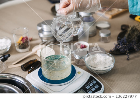 Hand wearing glove pours clear liquid from beaker into glass jar on digital scale. micro-batch beauty, small business production, handcrafted moisturizer, artisanal beauty lab. Hand wearing glove pours clear liquid from beaker into glass jar on digital scale. micro-batch beauty, small business production, handcrafted moisturizer, artisanal beauty lab. 136227977