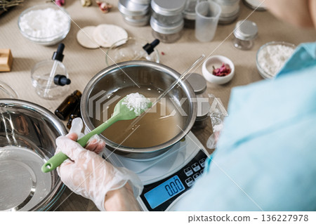 Person uses green spatula to hold white crystalline substance over metal bowl on digital scale. Hydration science aesthetic, pouring skincare liquid, water based cosmetic, refreshing beauty process. Person uses green spatula to hold white crystalline substance over metal bowl on digital scale. Hydration science aesthetic, pouring skincare liquid, water based cosmetic, refreshing beauty process. 136227978