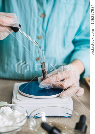 Gloved hands use glass dropper to add liquid to botanical ingredients in test tube on digital scale. Liquid boosters, floral droplets, herbal vials, custom blends. Gloved hands use glass dropper to add liquid to botanical ingredients in test tube on digital scale. Liquid boosters, floral droplets, herbal vials, custom blends. 136227984