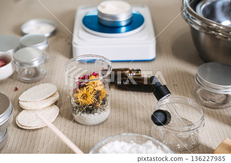 Beaker containing dried flowers and powder sits on work surface near cosmetic containers and scale. Raw beauty ingredients, skincare transparency, botanical extracts, minimalist cosmetic materials. Beaker containing dried flowers and powder sits on work surface near cosmetic containers and scale. Raw beauty ingredients, skincare transparency, botanical extracts, minimalist cosmetic materials. 136227985