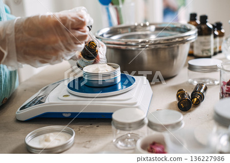 Person uses glass dropper to add oil to white cream in metal jar on digital scale. Lab-style beauty aesthetic, precise cosmetic formulation, diy apothecary lab, scientific skincare diy. Person uses glass dropper to add oil to white cream in metal jar on digital scale. Lab-style beauty aesthetic, precise cosmetic formulation, diy apothecary lab, scientific skincare diy. 136227986