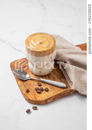 Dalgona coffee. Whipped instant iced coffee in a glass on a wooden board with coffee beans and spoon 136228928