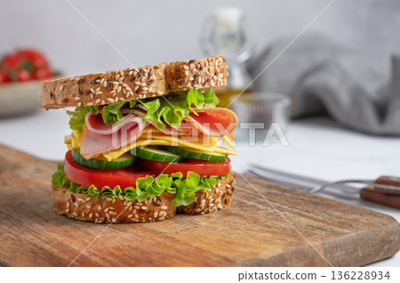Sandwich with ham, cheese, lettuce, tomatoes, cucumbers and seeded bread on a wooden board 136228934