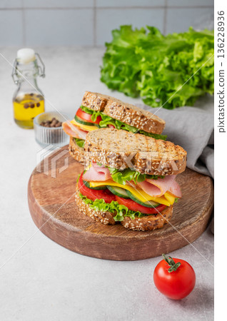 Two sandwiches with ham, cheese, lettuce and vegetable on a wooden board on a concrete background Two sandwiches with ham, cheese, lettuce and vegetable on a wooden board on a concrete background 136228936