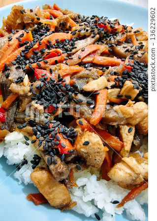 Chicken stir fry with sliced bell peppers and vegetables placed on white rice in a blue bowl. Black sesame seeds add texture on top of the dish Chicken stir fry with sliced bell peppers and vegetables placed on white rice in a blue bowl. Black sesame seeds add texture on top of the dish 136229602