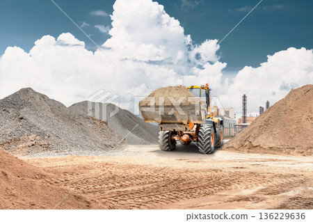 Loader picks up sand and transports it on a construction site surrounded by piles of earth and buildings in the background. Work is ongoing during daytime 136229636