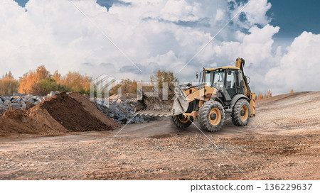 A wheel loader works at a construction site, moving soil and rock. The area is open, surrounded by piles of earth under a clear sky A wheel loader works at a construction site, moving soil and rock. The area is open, surrounded by piles of earth under a clear sky 136229637