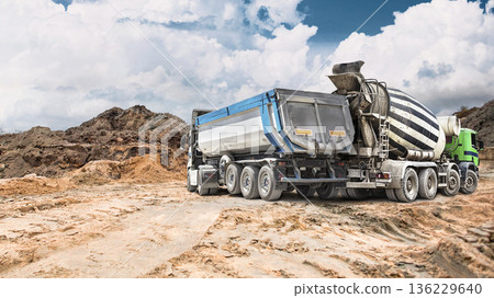 Construction equipment dump truck and concrete mixer truck at a construction site with earth embankments under a partly cloudy sky, with the focus on the heavy equipment Construction equipment dump truck and concrete mixer truck at a construction site with earth embankments under a partly cloudy sky, with the focus on the heavy equipment 136229640