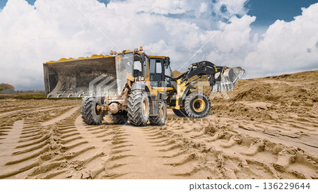 Deep tracks from machinery leave imprints on the sandy ground where two heavy loaders work under a cloudy sky. Rental equipment at a construction site 136229644