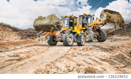 Two loaders carry sand and earth in buckets across a vast area of earthen banks, with clouds visible overhead Two loaders carry sand and earth in buckets across a vast area of earthen banks, with clouds visible overhead 136229652