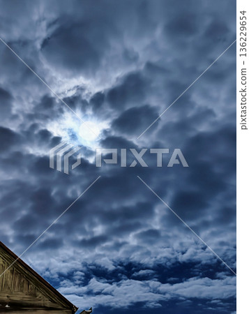 Puffy clouds fill the sky with a small area of sunlight breaking through. The scene depicts a late afternoon with a building in the foreground 136229654
