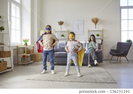 Kids in superhero customs having fun playing at home while their mother working on laptop Kids in superhero customs having fun playing at home while their mother working on laptop 136231191