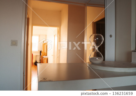 Hourglass on a white shelf with a sunlit apartment corridor in the background. Simple interior scene with natural light and everyday home objects. Hourglass on a white shelf with a sunlit apartment corridor in the background. Simple interior scene with natural light and everyday home objects. 136231659