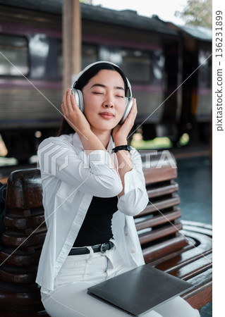 Woman in white shirt listening to music with headphones at a train station, sitting on a bench, eyes closed, enjoying a peaceful moment. 136231899