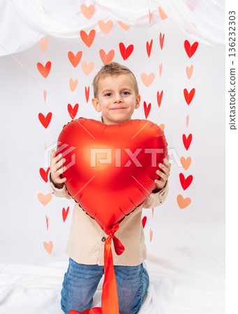 Happy little boy hugging big red heart balloon on white background. Valentines Day, love, warmth, emotional childhood portrait 136232303