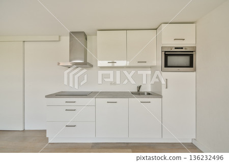 A sleek, modern kitchen featuring white cabinetry, a stainless steel range hood, and integrated appliances. The design emphasizes simplicity and functionality. 136232496