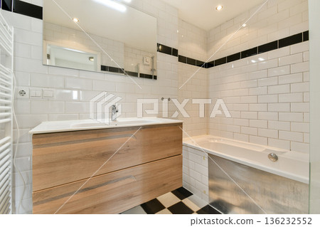 A sleek modern bathroom design featuring white tiled walls, a wooden vanity, and a bathtub. The space is illuminated with soft lighting for a cozy feel. A sleek modern bathroom design featuring white tiled walls, a wooden vanity, and a bathtub. The space is illuminated with soft lighting for a cozy feel. 136232552