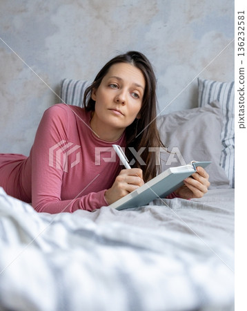 woman lies in bed writes in diary, writes emotions, wish list notebook. female writer plans workday. 136232581