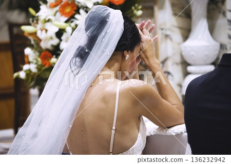View from behind a white Caucasian woman dressed as a bride in a white dress and veil on her head, kneeling and praying in a Catholic church at the altar 136232942