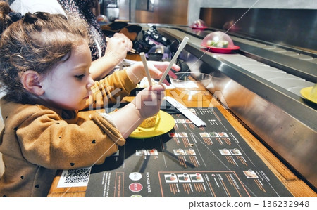Cute little girl using chopsticks to eat sushi at a restaurant with self-service conveyor belt 136232948