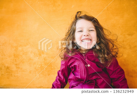 Young girl with a cheeky smile looking directly at the camera against a bright yellow wall background 136232949