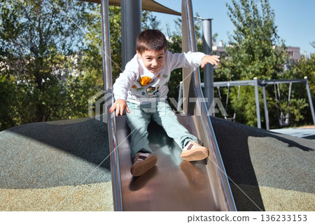 A white Caucasian boy smiling directly at the camera in an outdoor play park, sitting at the top of a slide A white Caucasian boy smiling directly at the camera in an outdoor play park, sitting at the top of a slide 136233153