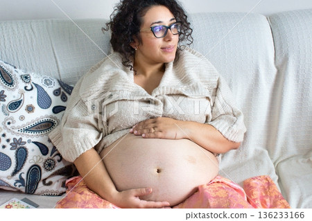 A young attractive white Caucasian woman sitting on a sofa at home, rubbing her pregnant belly, looking contemplative and thoughtful A young attractive white Caucasian woman sitting on a sofa at home, rubbing her pregnant belly, looking contemplative and thoughtful 136233166