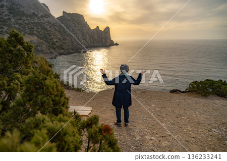 Woman cliff sunset view on rugged coastal mountains and shining sea expressing freedom 136233241