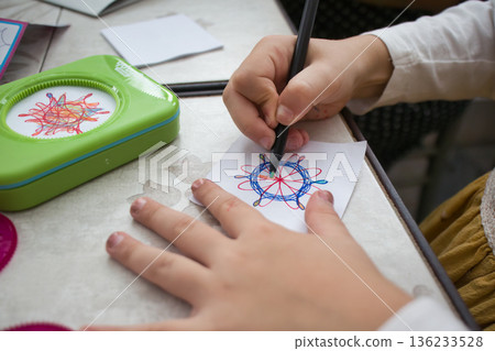 Close-up shot of the hands of a white Caucasian girl drawing a colorful spirograph design on paper 136233528
