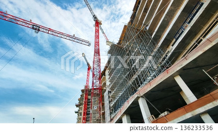 A ground-level worm's view of a construction site, looking up at scaffolding and cranes at the front of a building under construction 136233536