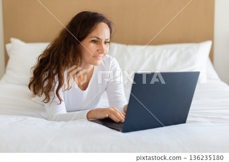 Middle aged woman using laptop while lying on bed at home 136235180