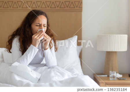 Ill adult caucasian woman sitting in bed blowing her nose with tissue 136235184