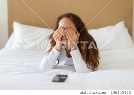Stressed woman lying on bed covering face while smartphone lies nearby Stressed woman lying on bed covering face while smartphone lies nearby 136235190