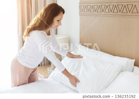 Adult caucasian woman making bed in bright bedroom, morning routine at home 136235192
