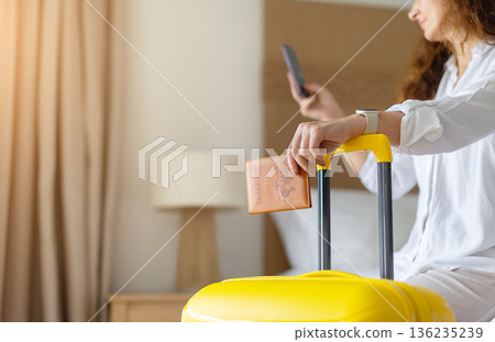 Adult caucasian woman at home holding passport and smartphone near suitcase 136235239