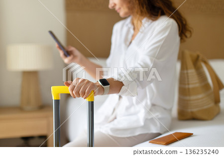 Adult caucasian woman holding suitcase handle while checking smartphone at home 136235240