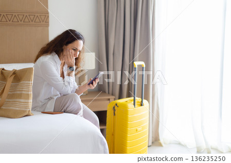 Woman sitting on bed at home, looking worried at smartphone beside suitcase Woman sitting on bed at home, looking worried at smartphone beside suitcase 136235250