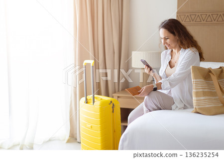Adult woman sitting on bed, using smartphone near suitcase, preparing for travel 136235254