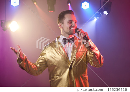 Happy showman in golden jacket singing into microphone on smoky party stage Happy showman in golden jacket singing into microphone on smoky party stage 136236516