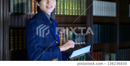 一位身穿西裝的年輕日本男子麵帶微笑，手拿平板電腦，在他的書房裡。 136236830