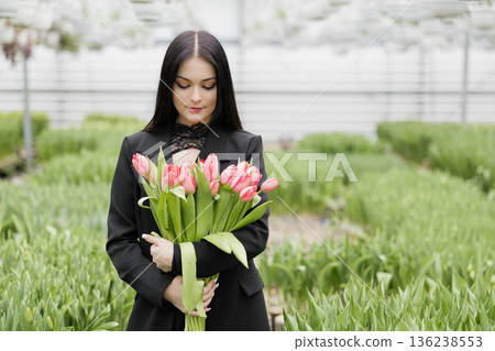 Young woman standing in large greenhouse and holding a blooming tulips in her hands Young woman standing in large greenhouse and holding a blooming tulips in her hands 136238553